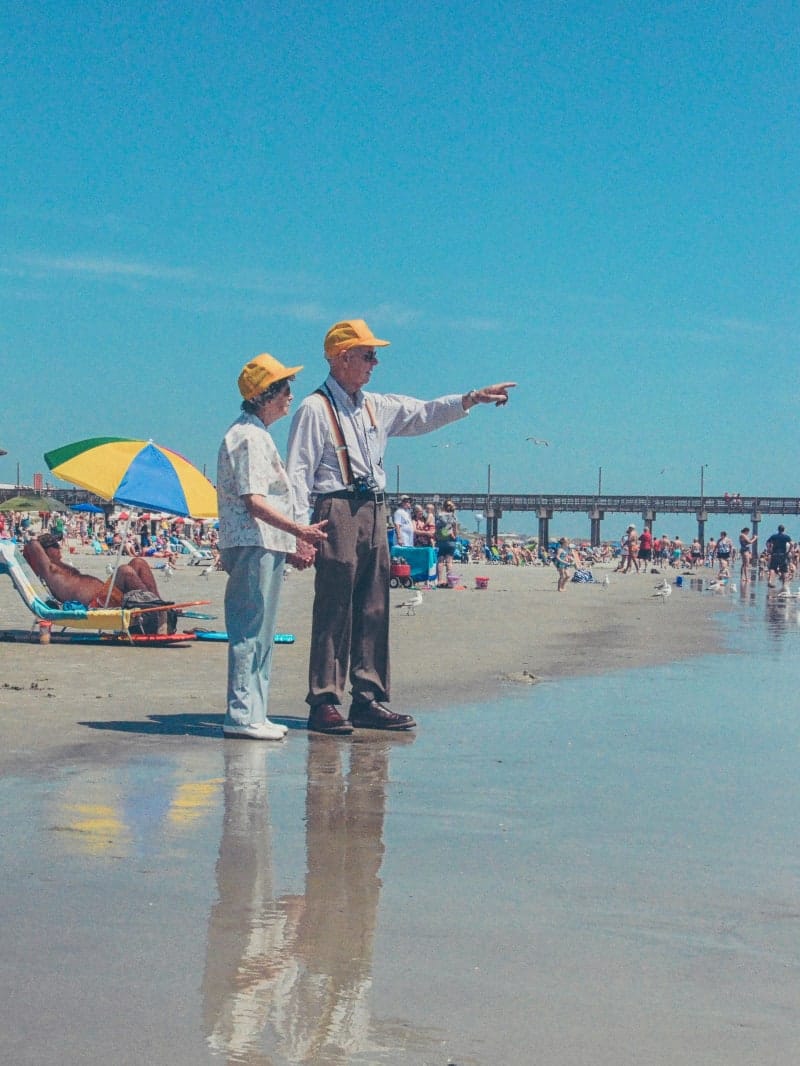 An old couple on a crowded beach, facing the sea. The man is pointing at the horizon.